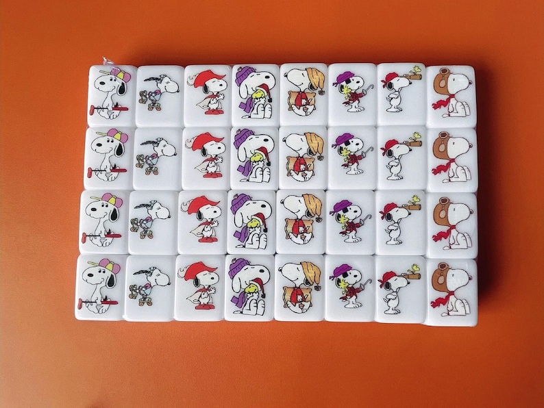 May include: A set of 36 white dominoes with colorful images of Snoopy and Woodstock. Each domino features a different image of the characters, including Snoopy wearing a hat, holding a bird, and wearing a scarf.