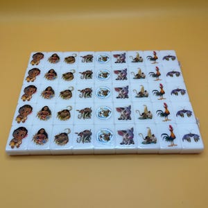 May include: A set of white Mahjong tiles featuring colorful illustrations of characters and symbols. The tiles are arranged in a rectangular shape on a yellow surface. The illustrations include a baby, a woman, a pig, a rooster, and other designs.