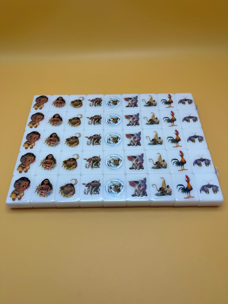 May include: A set of white Mahjong tiles featuring colorful illustrations of characters and symbols. The tiles are arranged in a rectangular shape on a yellow surface. The illustrations include a baby, a woman, a pig, a rooster, and other designs.