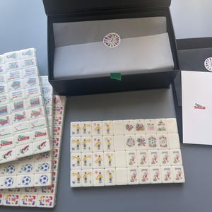 Puede incluir: Un juego de fichas de mahjong blancas con ilustraciones coloridas de tem&aacute;tica futbol&iacute;stica, que incluyen se&ntilde;ales de &aacute;rbitro, balones de f&uacute;tbol y jugadores. Incluye una caja de almacenamiento negra con papel seda, una tarjeta blanca y sobres negros.