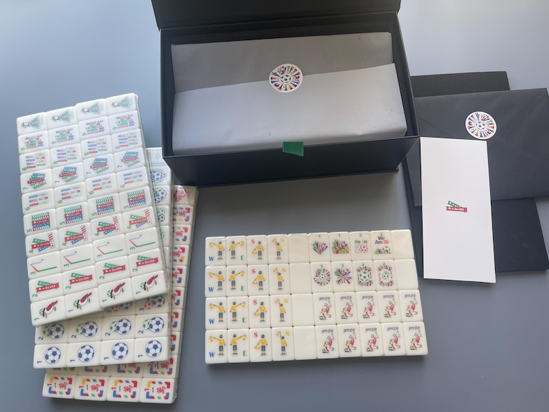 Puede incluir: Un juego de fichas de mahjong blancas con ilustraciones coloridas de tem&aacute;tica futbol&iacute;stica, que incluyen se&ntilde;ales de &aacute;rbitro, balones de f&uacute;tbol y jugadores. Incluye una caja de almacenamiento negra con papel seda, una tarjeta blanca y sobres negros.
