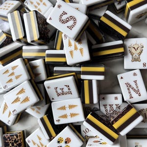 May include: A collection of rectangular mahjong tiles featuring white faces with gold, black, and brown printed designs including letters, numbers, pine trees, ladybugs, and leopard patterns. The sides of the tiles are layered with white, yellow, and black.