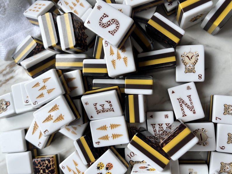 May include: A collection of rectangular mahjong tiles featuring white faces with gold, black, and brown printed designs including letters, numbers, pine trees, ladybugs, and leopard patterns. The sides of the tiles are layered with white, yellow, and black.