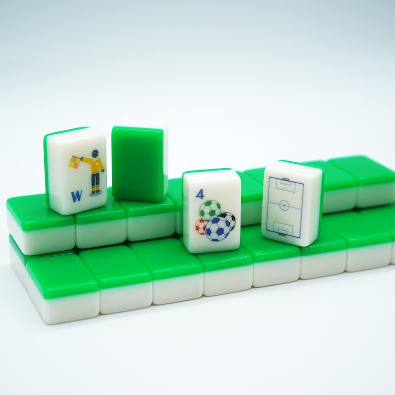 Puede incluir: Una colecci&oacute;n de fichas de mahjong rectangulares con reverso verde y frente blanco. Tres fichas est&aacute;n de pie, mostrando un &aacute;rbitro con una bandera y la letra W, el n&uacute;mero 4 con balones de f&uacute;tbol y el contorno azul de un campo de f&uacute;tbol.
