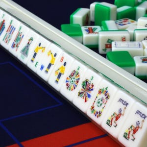 Puede incluir: Un conjunto de fichas de Mahjong blancas y verdes dispuestas en un soporte y esparcidas sobre una superficie. Las fichas presentan ilustraciones coloridas de figuras deportivas, banderas y balones, con inscripciones JOKER, E, W y S.