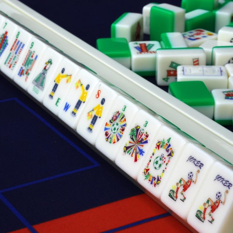Puede incluir: Un conjunto de fichas de Mahjong blancas y verdes dispuestas en un soporte y esparcidas sobre una superficie. Las fichas presentan ilustraciones coloridas de figuras deportivas, banderas y balones, con inscripciones JOKER, E, W y S.