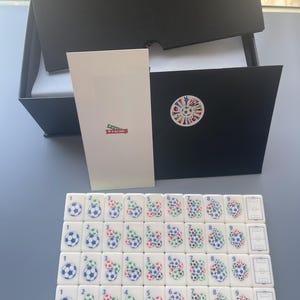 Puede incluir: Un conjunto de fichas rectangulares blancas dispuestas en cuatro filas, cada una con gr&aacute;ficos de balones de f&uacute;tbol y n&uacute;meros del uno al nueve, junto a una caja negra, una tarjeta blanca con una banda roja que dice Go USA y un sobre negro con pegatina.