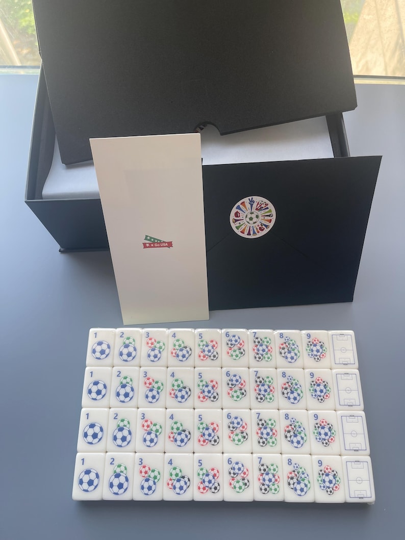 Puede incluir: Un conjunto de fichas rectangulares blancas dispuestas en cuatro filas, cada una con gr&aacute;ficos de balones de f&uacute;tbol y n&uacute;meros del uno al nueve, junto a una caja negra, una tarjeta blanca con una banda roja que dice Go USA y un sobre negro con pegatina.