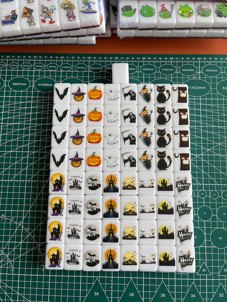 May include: A set of 36 white ceramic tiles with Halloween-themed designs. The tiles feature images of pumpkins, ghosts, bats, witches, and castles. The tiles are arranged in a grid pattern and are ready to be used for a game of mahjong or other tile-based games.