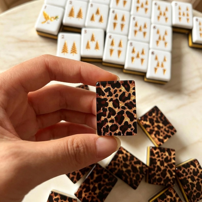 May include: A rectangular mahjong tile featuring a brown and black leopard print pattern with a gold glitter finish. The tile is held upright, while other matching leopard print tiles and white tiles with gold tree designs are scattered nearby.