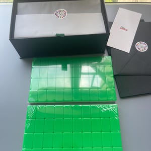 Puede incluir: Una caja de regalo negra que contiene papel de seda blanco con una pegatina circular de banderas coloridas, dos juegos rectangulares de fichas de mahjong de color verde brillante envueltas en pl&aacute;stico, una tarjeta blanca y un sobre negro.