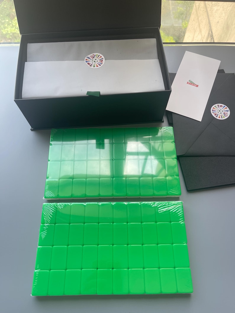 Puede incluir: Una caja de regalo negra que contiene papel de seda blanco con una pegatina circular de banderas coloridas, dos juegos rectangulares de fichas de mahjong de color verde brillante envueltas en pl&aacute;stico, una tarjeta blanca y un sobre negro.