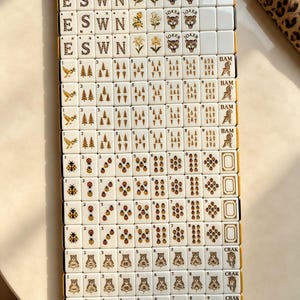 May include: A set of cream-colored Mahjong tiles arranged in a grid. The tiles feature gold-toned illustrations including letters E, S, W, N, floral motifs, tiger faces labeled Joker, birds, pine trees, ladybugs, and tiger figures with numbers.