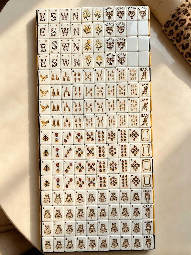 May include: A set of cream-colored Mahjong tiles arranged in a grid. The tiles feature gold-toned illustrations including letters E, S, W, N, floral motifs, tiger faces labeled Joker, birds, pine trees, ladybugs, and tiger figures with numbers.
