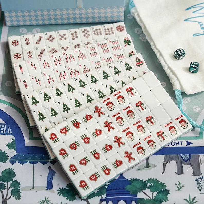 Op de afbeelding: Een set witte Mahjong-tegels met kerstthema-illustraties zoals de Kerstman, peperkoekmannetjes, kerstbomen, cadeautjes en zuurstokken, inclusief twee groene dobbelstenen en een wit opbergzakje met trekkoord en blauwe tekst.