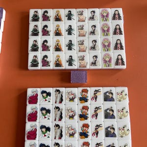 May include: White tiles with colorful anime character illustrations, arranged in sets. The tiles feature characters from popular anime series. A small, glittery purple tile is also present. The tiles are on an orange surface.
