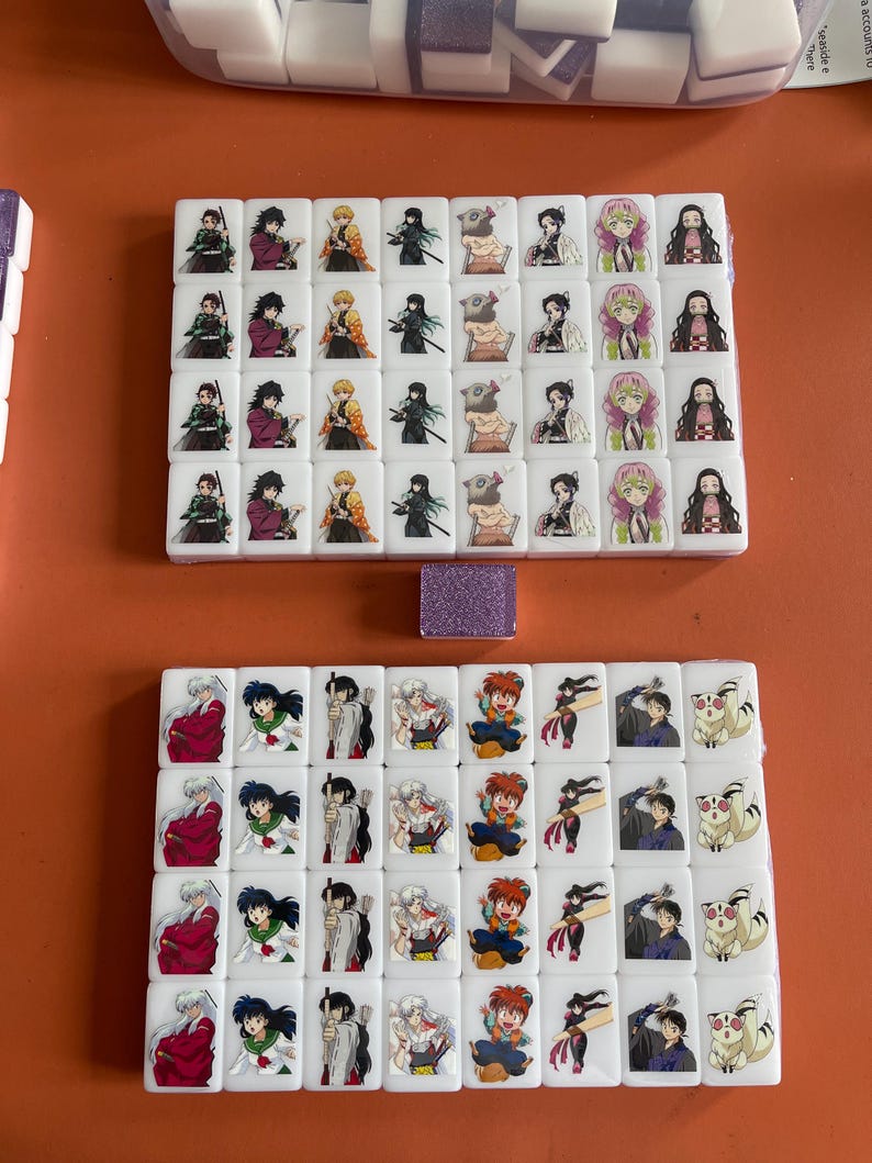 May include: White tiles with colorful anime character illustrations, arranged in sets. The tiles feature characters from popular anime series. A small, glittery purple tile is also present. The tiles are on an orange surface.
