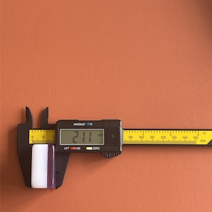 May include: A digital caliper measuring a rectangular object. The object is white with a purple stripe and measures 21.1 mm, or approximately 0.83 inches. The caliper is black with a yellow and black ruler.