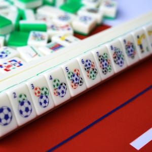 Puede incluir: Un conjunto de fichas de mahjong blancas con reverso verde, dispuestas en fila y en mont&oacute;n. Cada ficha presenta un n&uacute;mero azul y un gr&aacute;fico de balones de f&uacute;tbol de varios colores sobre una superficie roja con una l&iacute;nea azul fina.