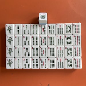 May include: A set of white Mahjong tiles arranged on an orange surface. The tiles display various symbols, numbers, and characters, including the word "JOKER" on a single tile. The game pieces are for a traditional tile-based game.