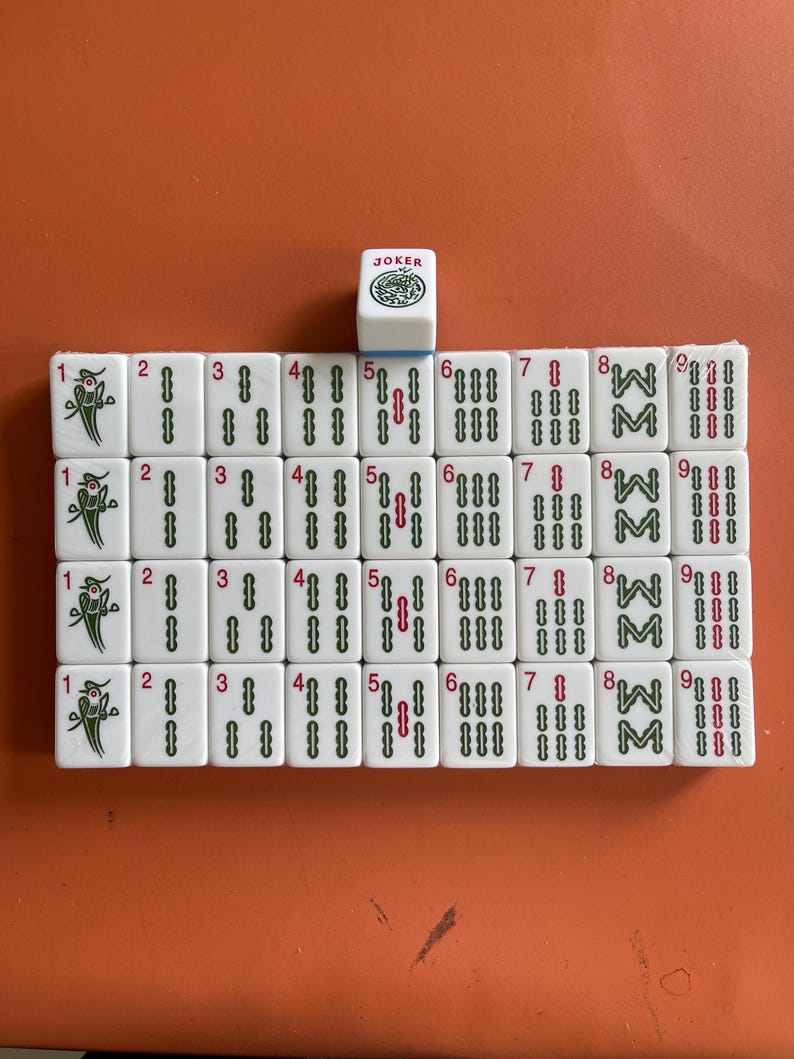 May include: A set of white Mahjong tiles arranged on an orange surface. The tiles display various symbols, numbers, and characters, including the word "JOKER" on a single tile. The game pieces are for a traditional tile-based game.