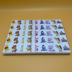 May include: A set of white mahjong tiles featuring colorful illustrations of Winnie the Pooh and friends. Each tile displays a different character, including Pooh, Tigger, Piglet, Eeyore, and others. The tiles are arranged in a rectangular pattern.