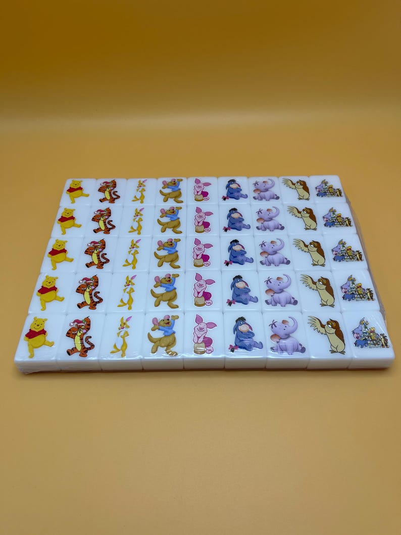 May include: A set of white mahjong tiles featuring colorful illustrations of Winnie the Pooh and friends. Each tile displays a different character, including Pooh, Tigger, Piglet, Eeyore, and others. The tiles are arranged in a rectangular pattern.