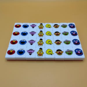 May include: A set of white tiles featuring colorful Sesame Street character faces. The tiles are arranged in rows and columns, showcasing characters like Elmo, Cookie Monster, and Bert and Ernie. The tiles are likely part of a game.