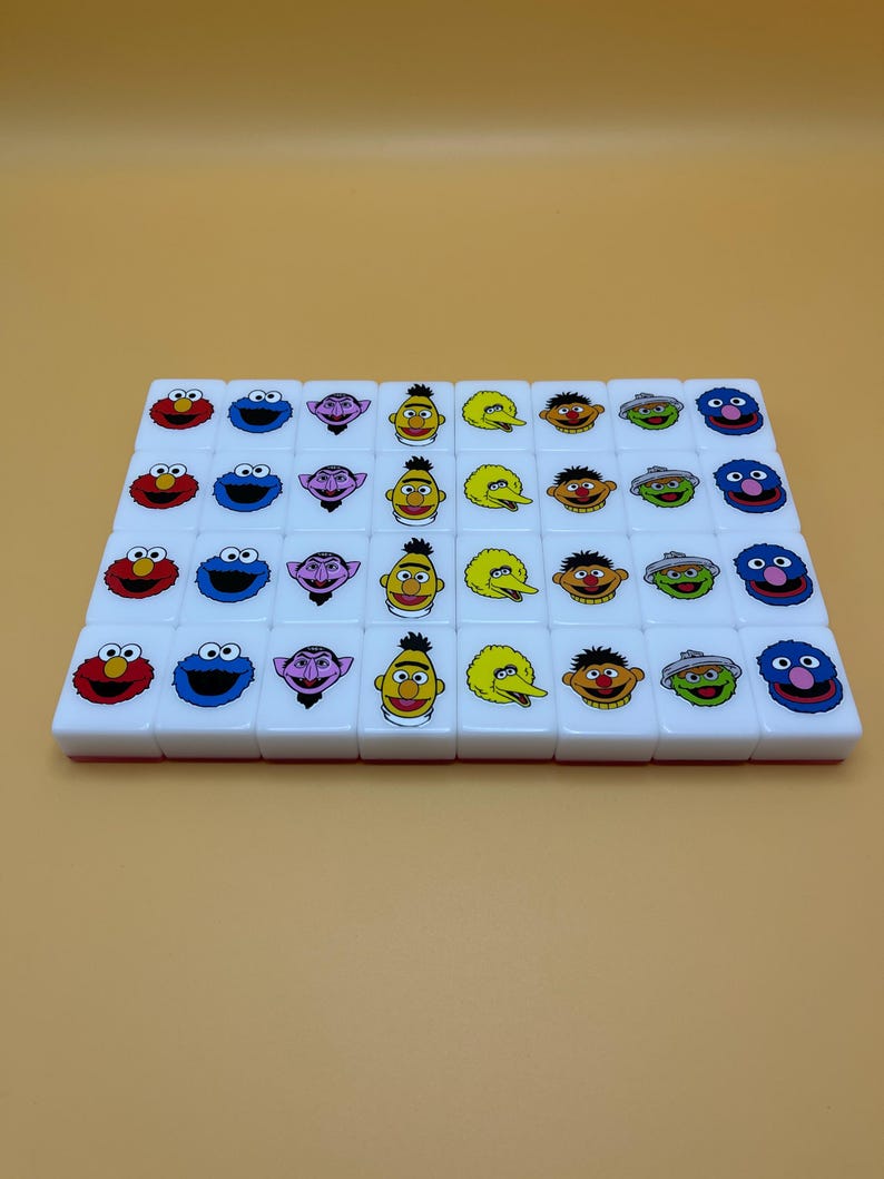 May include: A set of white tiles featuring colorful Sesame Street character faces. The tiles are arranged in rows and columns, showcasing characters like Elmo, Cookie Monster, and Bert and Ernie. The tiles are likely part of a game.
