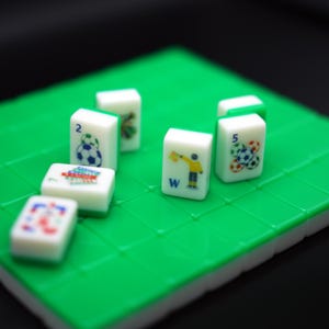Puede incluir: Un conjunto de fichas rectangulares blancas con gr&aacute;ficos coloridos de tem&aacute;tica deportiva, incluyendo balones de f&uacute;tbol y un &aacute;rbitro, dispuestas sobre un tablero de rejilla verde brillante. Las fichas muestran n&uacute;meros y letras, concretamente el 2, el 5 y la W.