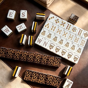 May include: A Mahjong set featuring white tiles with leopard print backs and sides with black and yellow stripes. The tiles display various illustrations including tigers, ladybugs, pine trees, and the word JOKER, along with letters E, S, M, Z, and BAM.