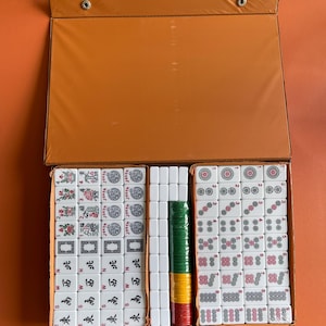 May include: A set of white mahjong tiles in a brown case. The tiles are arranged in rows and columns, with the numbers 1 through 9 visible on the tiles. The case has a flap that opens to reveal the tiles.