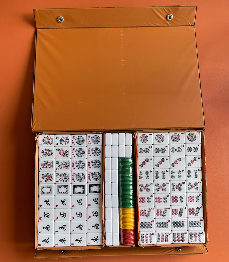 May include: A set of white mahjong tiles in a brown case. The tiles are arranged in rows and columns, with the numbers 1 through 9 visible on the tiles. The case has a flap that opens to reveal the tiles.