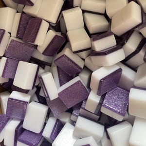 May include: A close-up shot of a pile of white and purple Mahjong tiles. The tiles are rectangular with white sides and purple tops. The purple tops have a textured, glittery appearance. The tiles are scattered, creating a full-frame image.