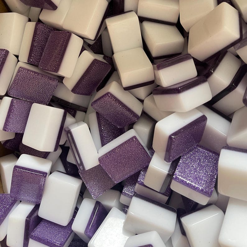 May include: A close-up shot of a pile of white and purple Mahjong tiles. The tiles are rectangular with white sides and purple tops. The purple tops have a textured, glittery appearance. The tiles are scattered, creating a full-frame image.