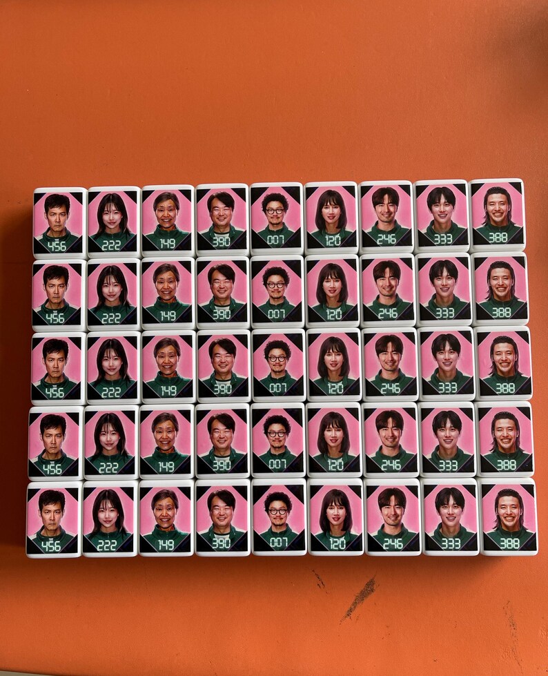May include: A set of 35 white tiles with pink backgrounds and black numbers. Each tile features a different person's face. The numbers on the tiles are 456, 222, 149, 390, 007, 120, 246, 333, and 388.