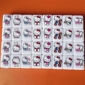May include: A set of 36 white dominoes with Hello Kitty themed illustrations. Each domino features a different Hello Kitty design, including Hello Kitty taking pictures, riding a bike, baking a cake, and painting.