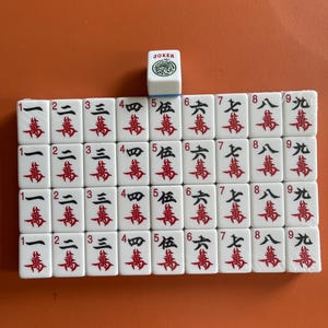 May include: A set of white Mahjong tiles with red and black Chinese characters and numbers. A single Joker tile is on top. The tiles are arranged on an orange surface.