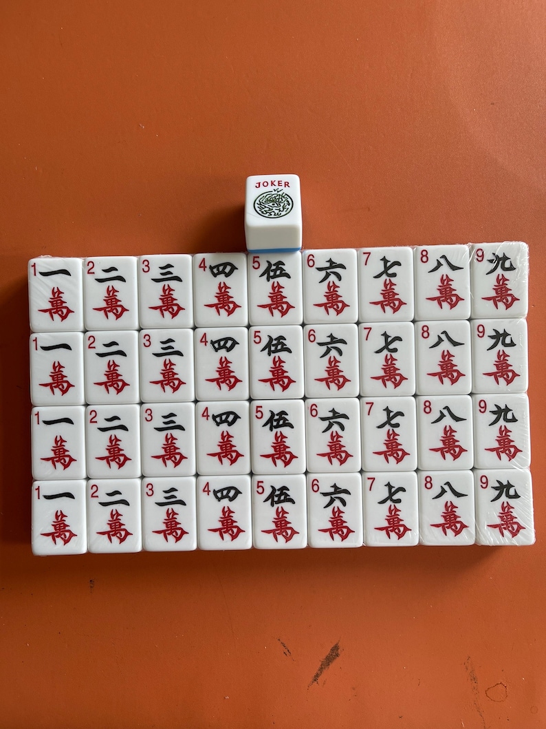 May include: A set of white Mahjong tiles with red and black Chinese characters and numbers. A single Joker tile is on top. The tiles are arranged on an orange surface.
