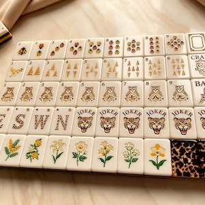 May include: A set of cream-colored Mahjong tiles arranged in a grid. The tiles feature gold-toned illustrations of ladybugs, trees, leopards, and flowers, along with the text JOKER, CRAK, BAM, E, S, W, N, and leopard print patterns on the final two tiles.