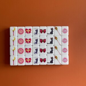 May include: A set of 24 white dominoes with images of characters from the anime Sailor Moon. The dominoes feature a crescent moon, a star, a heart, a cat, a bow, a wand, and a lotus flower.