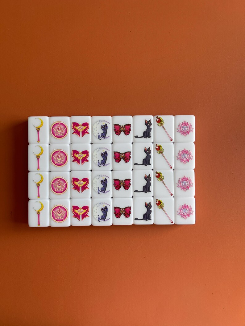 May include: A set of 24 white dominoes with images of characters from the anime Sailor Moon. The dominoes feature a crescent moon, a star, a heart, a cat, a bow, a wand, and a lotus flower.