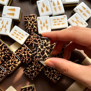May include: A collection of rectangular mahjong tiles featuring two distinct designs. Some tiles are white with gold-colored pine tree illustrations and black numbers, while others display a brown and black leopard print pattern on the face.
