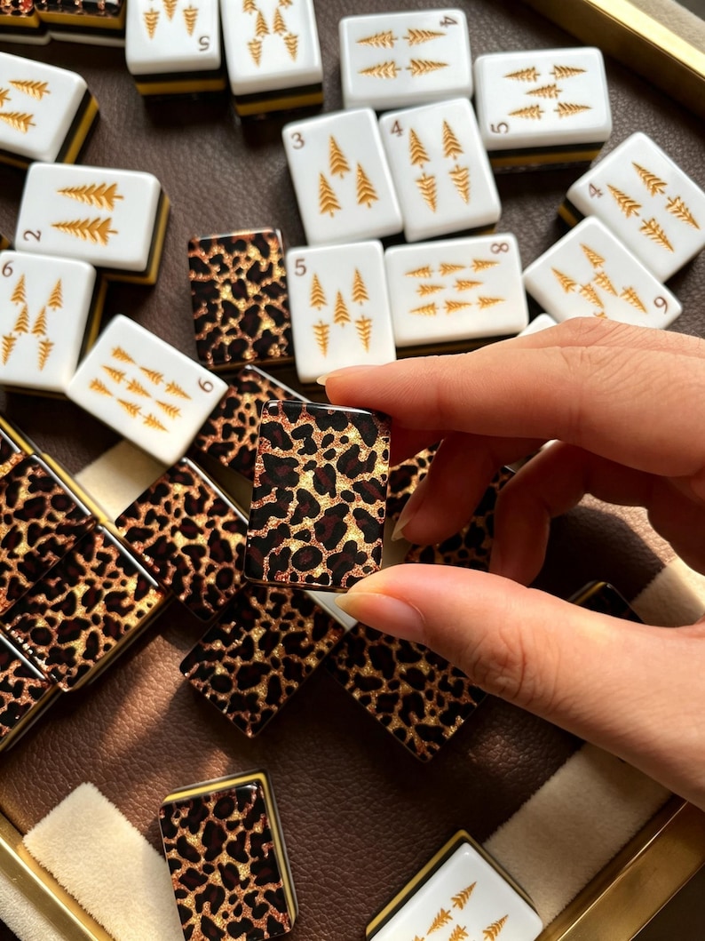 May include: A collection of rectangular mahjong tiles featuring two distinct designs. Some tiles are white with gold-colored pine tree illustrations and black numbers, while others display a brown and black leopard print pattern on the face.
