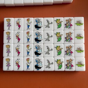 May include: A set of white mahjong tiles featuring cartoon characters. The tiles display colorful illustrations of characters in various poses. The tiles are arranged in rows, with some tiles stacked on top.