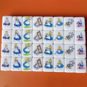 Op de afbeelding: Een set van 24 witte domino's met kleurrijke illustraties van Alice in Wonderland-personages. De domino's tonen Alice, de Cheshire Cat, de Hoedenmaker en de Koningin van harten.