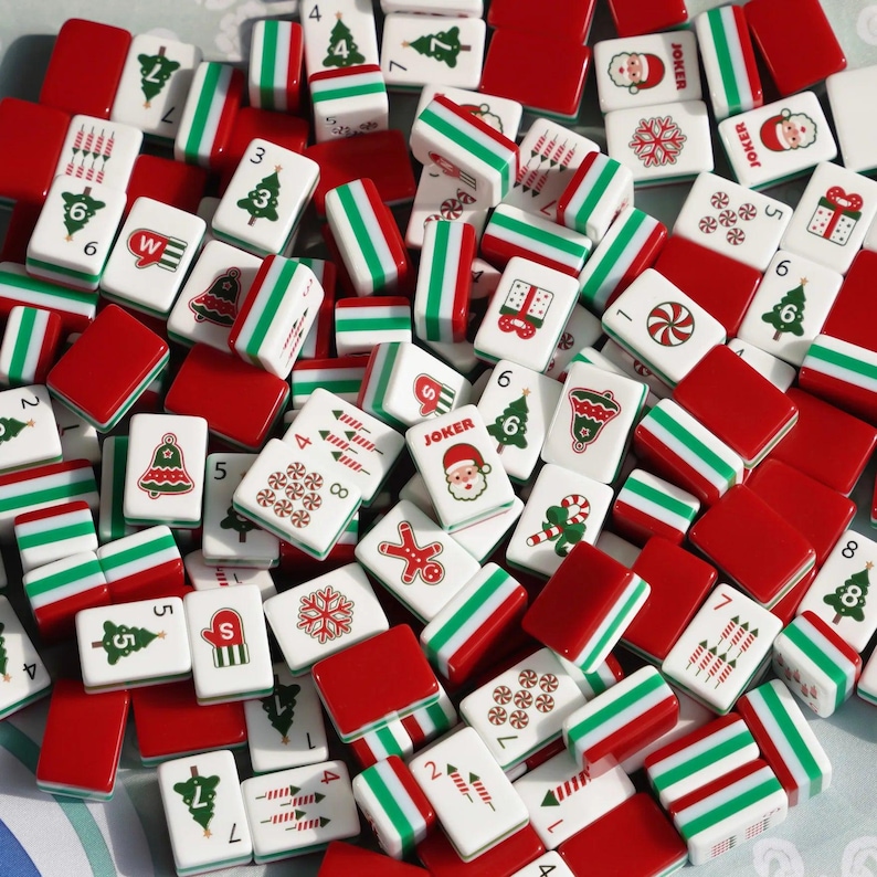 Op de afbeelding: Een verzameling rechthoekige mahjongtegels met kerstthema-illustraties, waaronder de Kerstman, versierde dennenbomen, klokken, sneeuwvlokken, zuurstokken, geschenkdozen en peperkoekmannetjes, afgewisseld met effen rode en groen-wit gestreepte tegels.