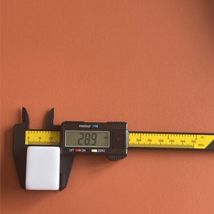 May include: A white rectangular object is measured with a black digital caliper. The caliper displays a measurement of 28.9 mm. The caliper has both mm and inch scales. The background is a solid orange-brown color.