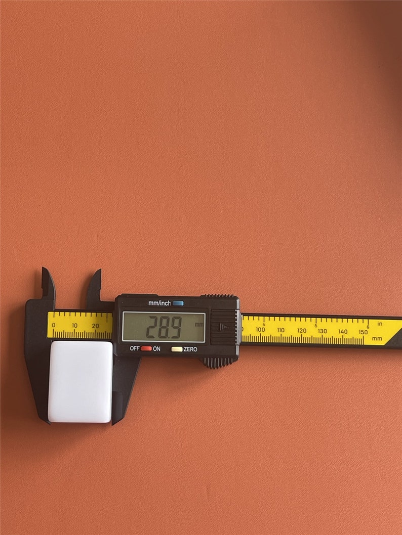 May include: A white rectangular object is measured with a black digital caliper. The caliper displays a measurement of 28.9 mm. The caliper has both mm and inch scales. The background is a solid orange-brown color.