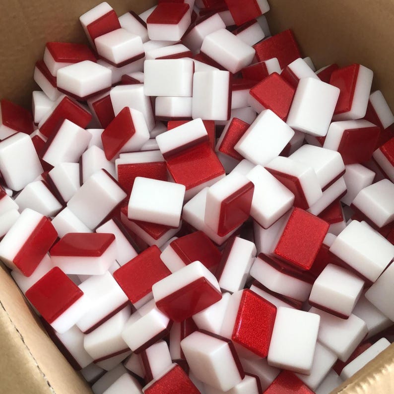 May include: A collection of white and red tiles, likely for a game like Mahjong or dominoes, fills a cardboard box. The tiles are rectangular, with a white surface and a red, possibly glittery, accent on one side. The image is a close-up, showing the tiles in a jumbled arrangement.